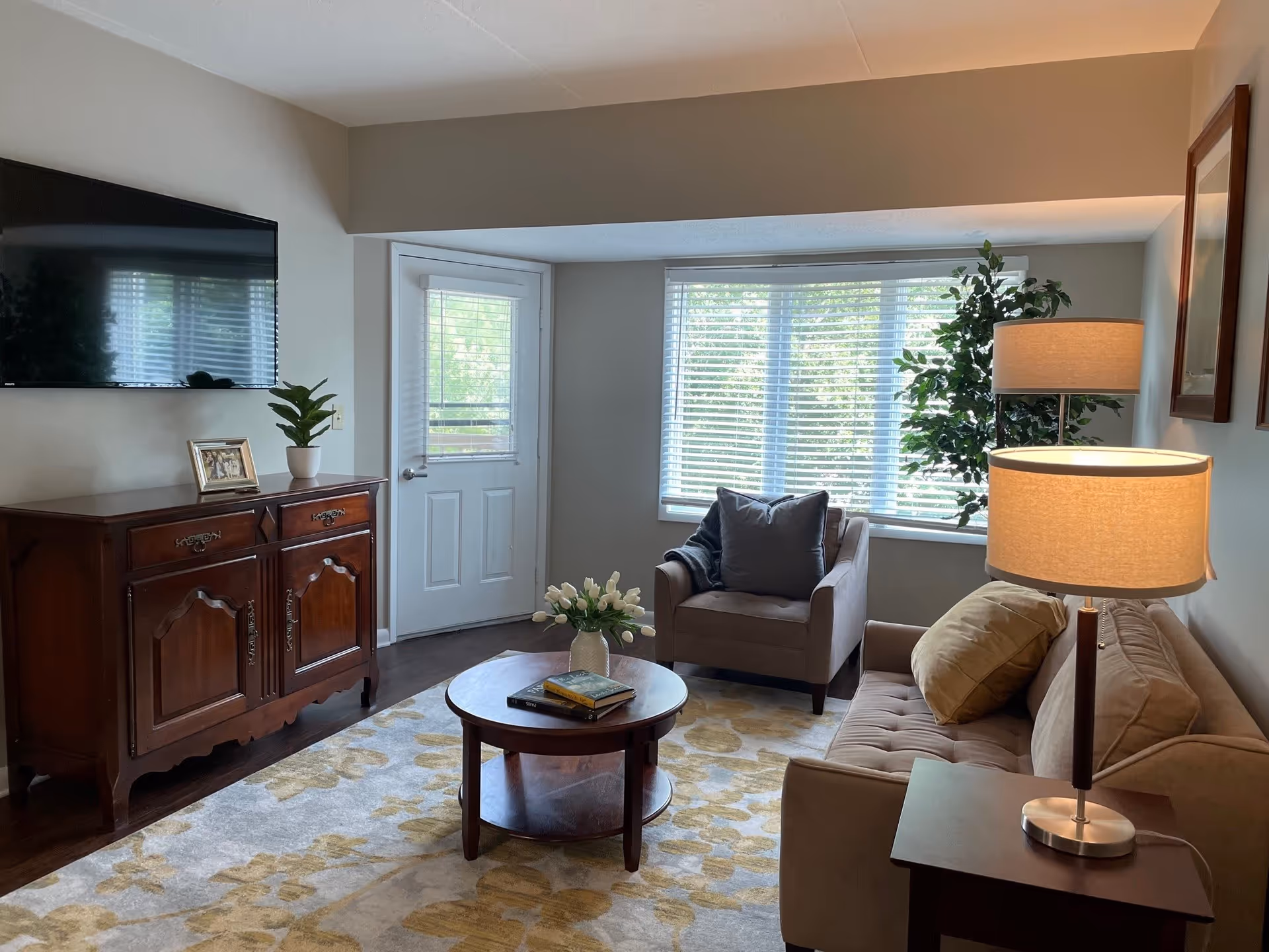 A cozy living room with a beige couch and matching armchair, a wooden coffee table with books and a vase of white tulips, a wooden cabinet with a potted plant and framed photo, a flat-screen TV mounted on the wall, a large window with blinds, a door with a window, and a floor lamp next to the couch.