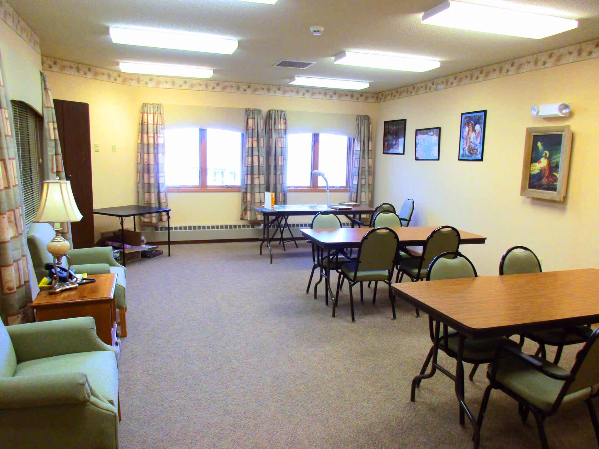 A well-lit room with beige walls and carpeted floor featuring several tables and chairs arranged for group seating. There are three windows with patterned curtains letting in natural light. On the left side, there are two green armchairs and a wooden side table with a lamp. The walls have framed pictures and a decorative border near the ceiling.