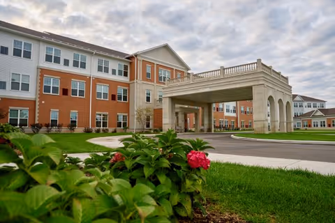 Exterior view of a large senior living facility building with a covered entrance driveway. The building has multiple windows and a combination of brick and light-colored siding. In the foreground, there are green plants with pink flowers and a well-maintained lawn under a cloudy sky.