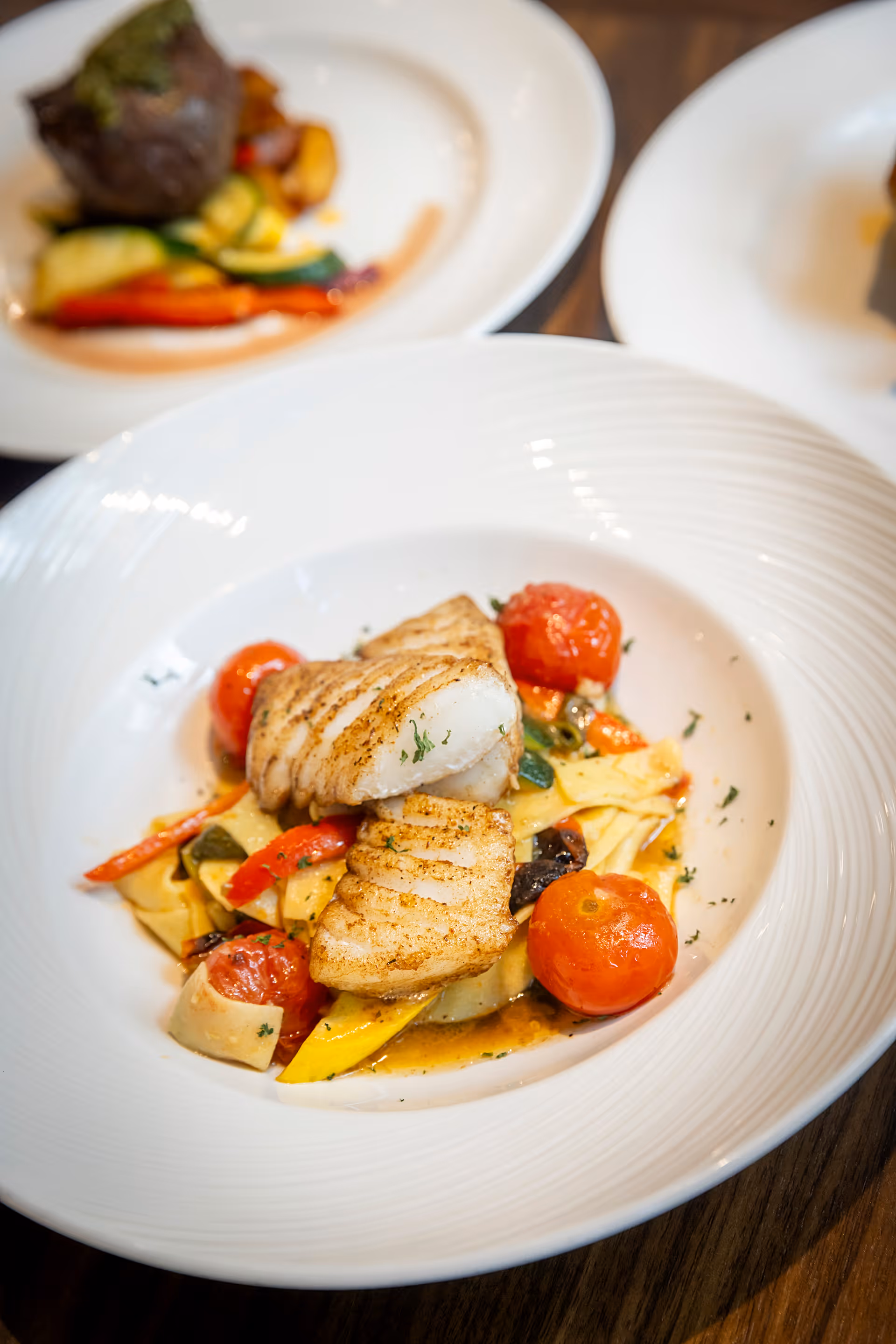 A white plate with grilled fish fillets served on a bed of mixed sautéed vegetables including cherry tomatoes, red bell peppers, and zucchini. In the background, there is another plate with a piece of meat and vegetables, slightly out of focus.