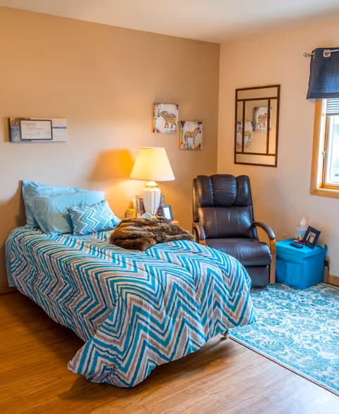 Single bedroom with a teal chevron-patterned bed, bedside lamp, leather recliner, and a window with a blue valance.