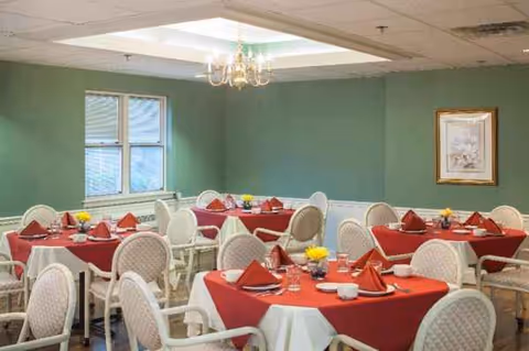 Dining room with red tablecloths, white chairs, and chandelier lighting in a facility.