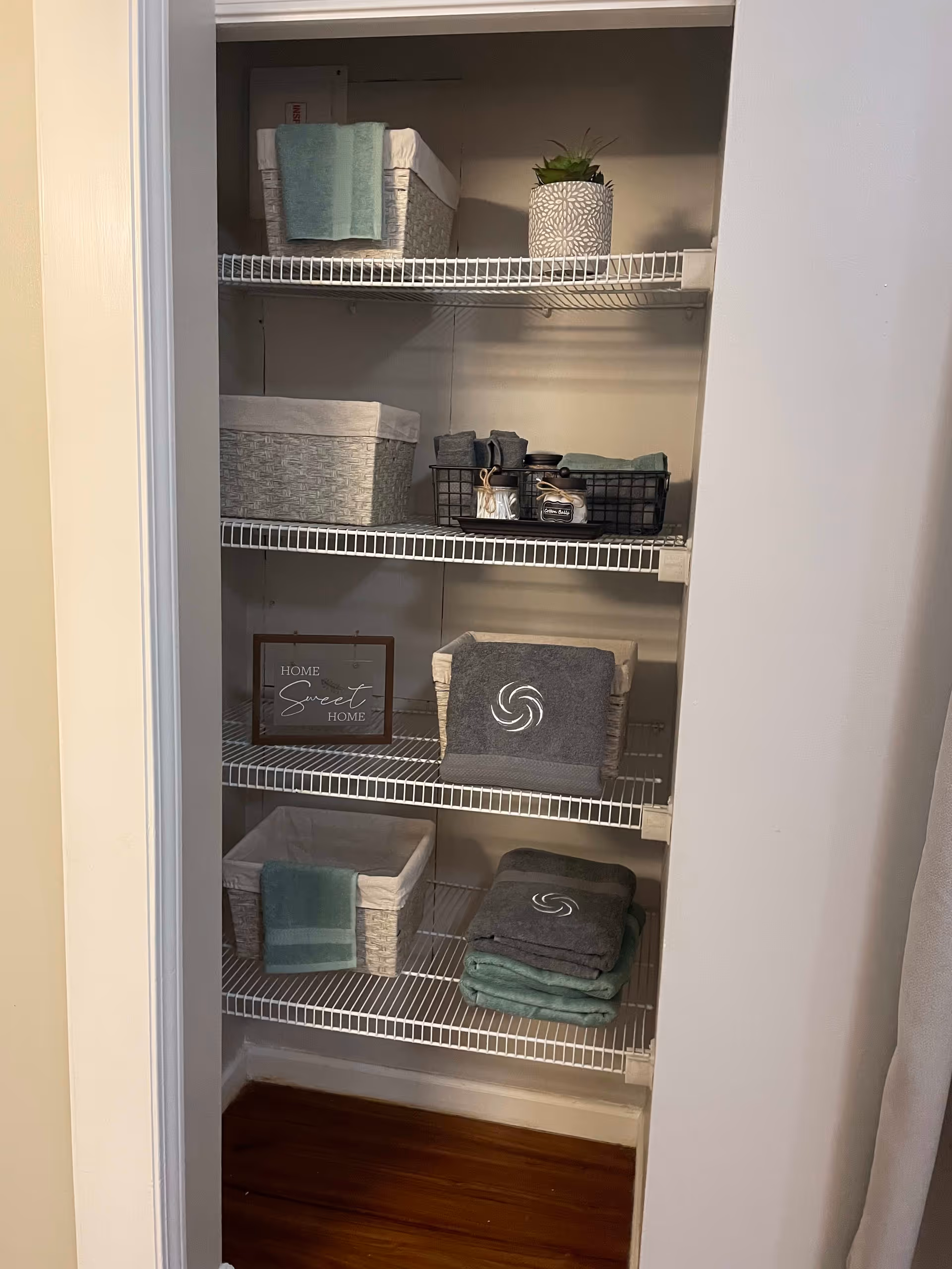 Open shelving unit with four wire shelves holding woven baskets, folded towels, a small potted plant, and a framed sign that reads 'Home Sweet Home'. The shelves are neatly organized and the floor beneath is wooden.