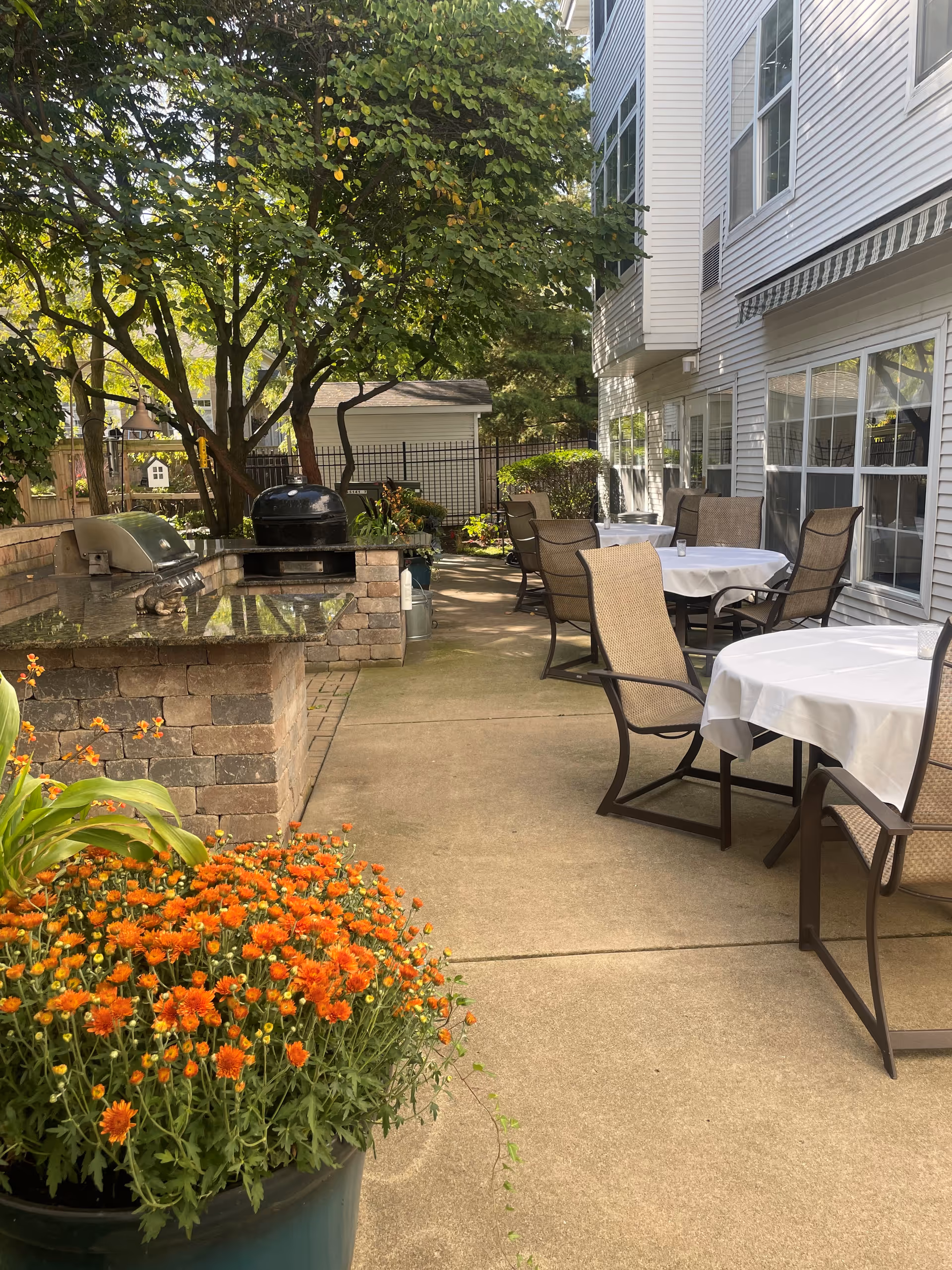 Outdoor patio area with several tables covered with white tablecloths and surrounded by chairs. There is a built-in brick grill and a black charcoal grill on the left side. The patio is next to a white building with multiple windows and an awning. A large potted plant with orange flowers is in the foreground, and trees provide shade in the background.