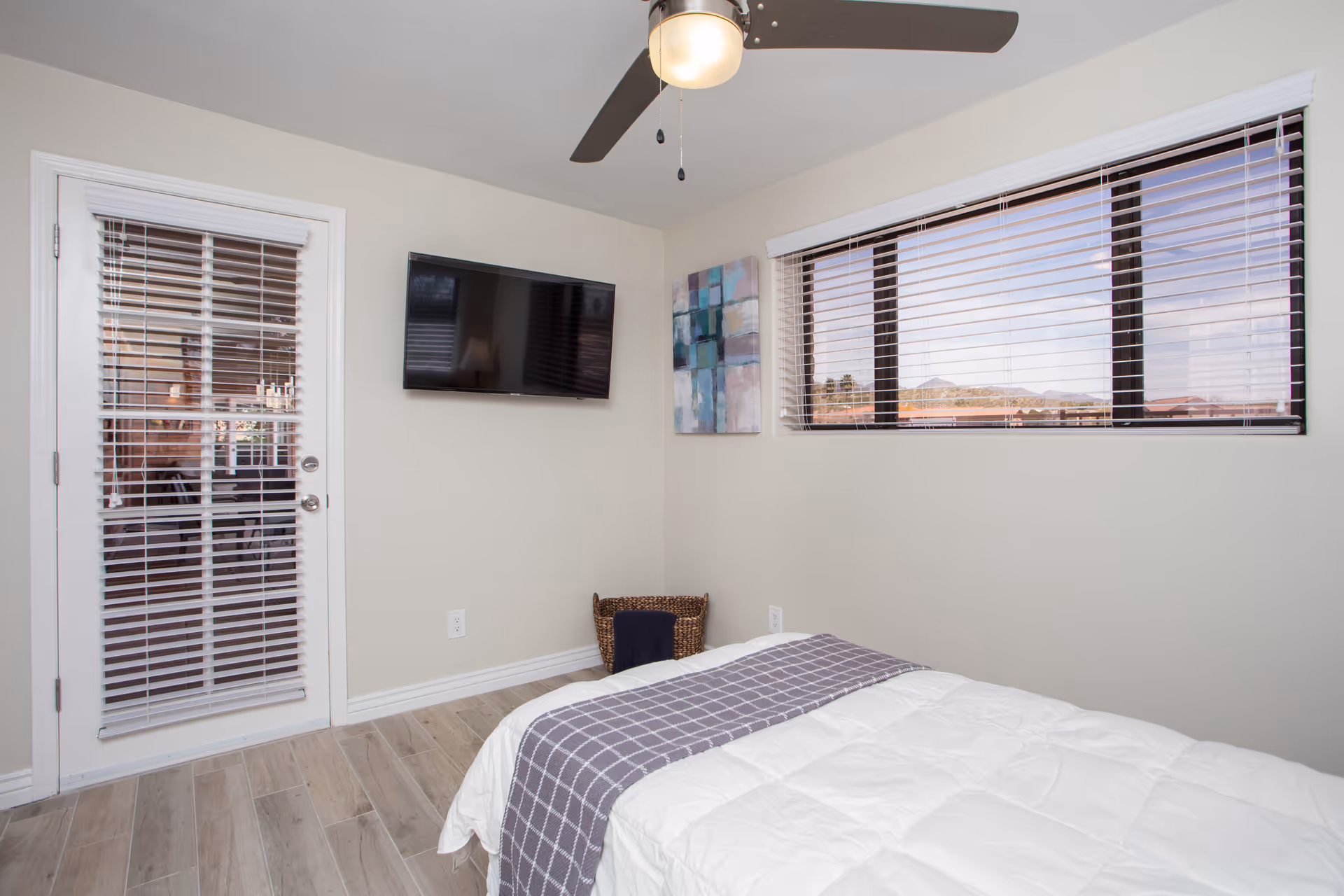 A bedroom with a bed covered in a white comforter and a gray checkered throw blanket. There is a ceiling fan with a light, a wall-mounted flat-screen TV, a window with blinds showing a view of mountains, a door with blinds, a wicker basket in the corner, and a colorful abstract painting on the wall.