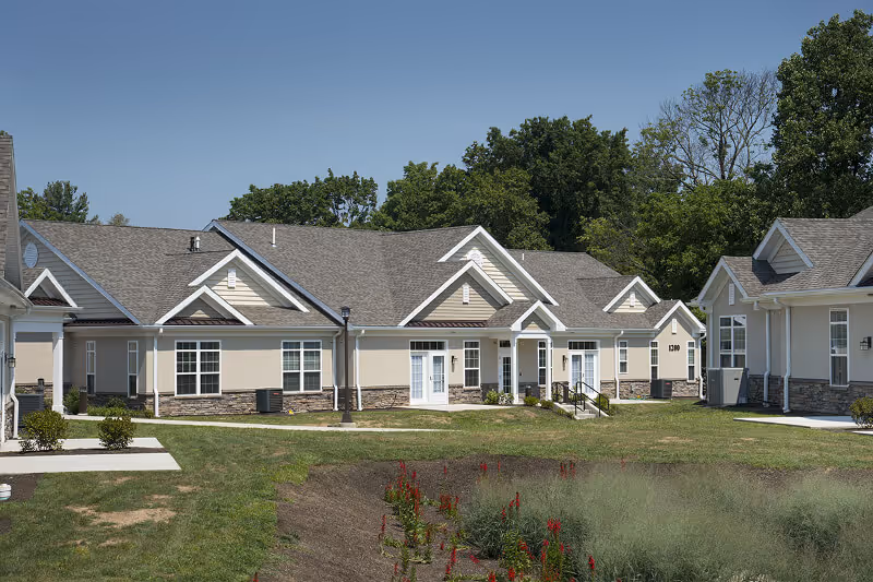 Exterior view of a single-story senior living facility building with beige walls, stone accents, multiple white-framed windows and doors, and a gray shingled roof. The building is surrounded by a grassy lawn with some small bushes and red flowers in the foreground, under a clear blue sky.
