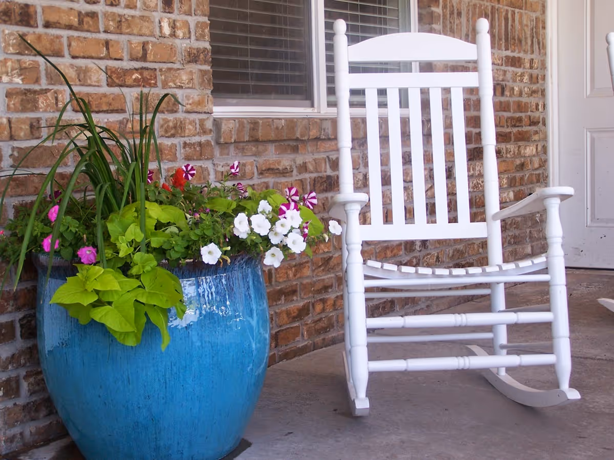 A white wooden rocking chair sits on a porch beside a large blue planter filled with green foliage and colorful flowers against a brick wall.