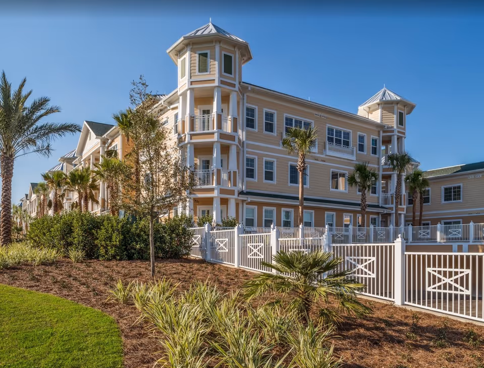 Three-story pale yellow senior living building with wraparound balconies, palm trees, and a white fence under a clear blue sky.