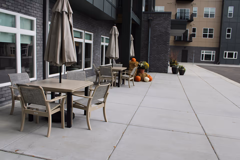 Outdoor patio area with several tables and chairs, each table having a closed umbrella. The patio is adjacent to a building with multiple windows. There are fall decorations including pumpkins and potted flowers near the building wall.