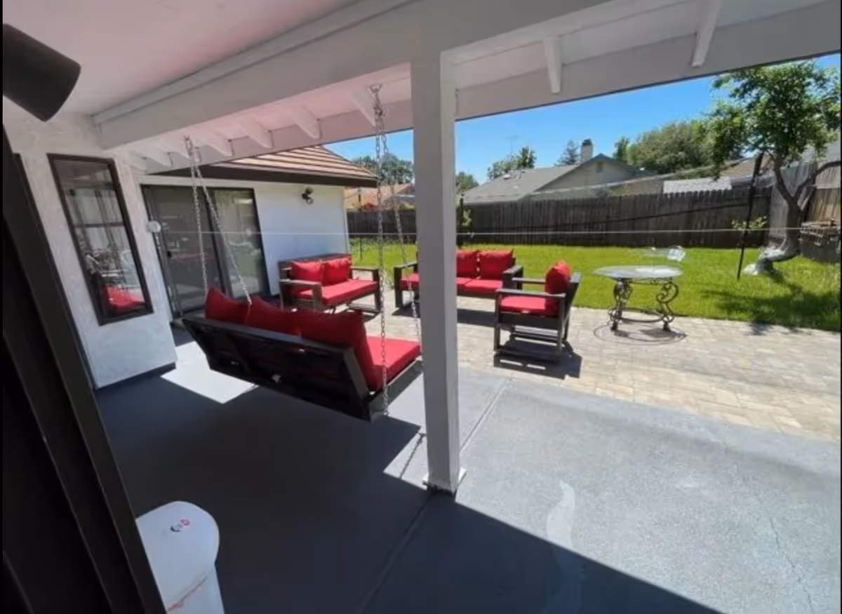 Covered patio with red-cushioned outdoor seating, a hanging porch swing, and a grassy backyard with a fence.