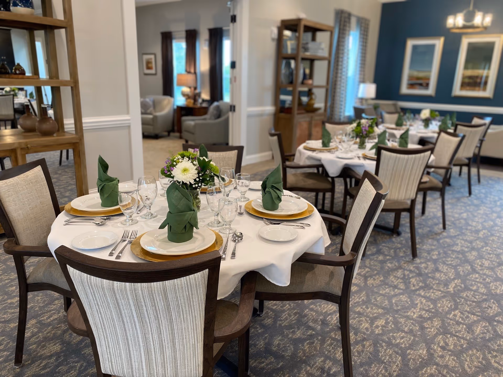 A dining room in an assisted living facility with round tables set for a meal. Each table has white tablecloths, green folded napkins, plates, glasses, silverware, and floral centerpieces. The room has carpeted floors, upholstered chairs, and blue accent walls with framed artwork. In the background, there is a seating area with armchairs and a lamp.