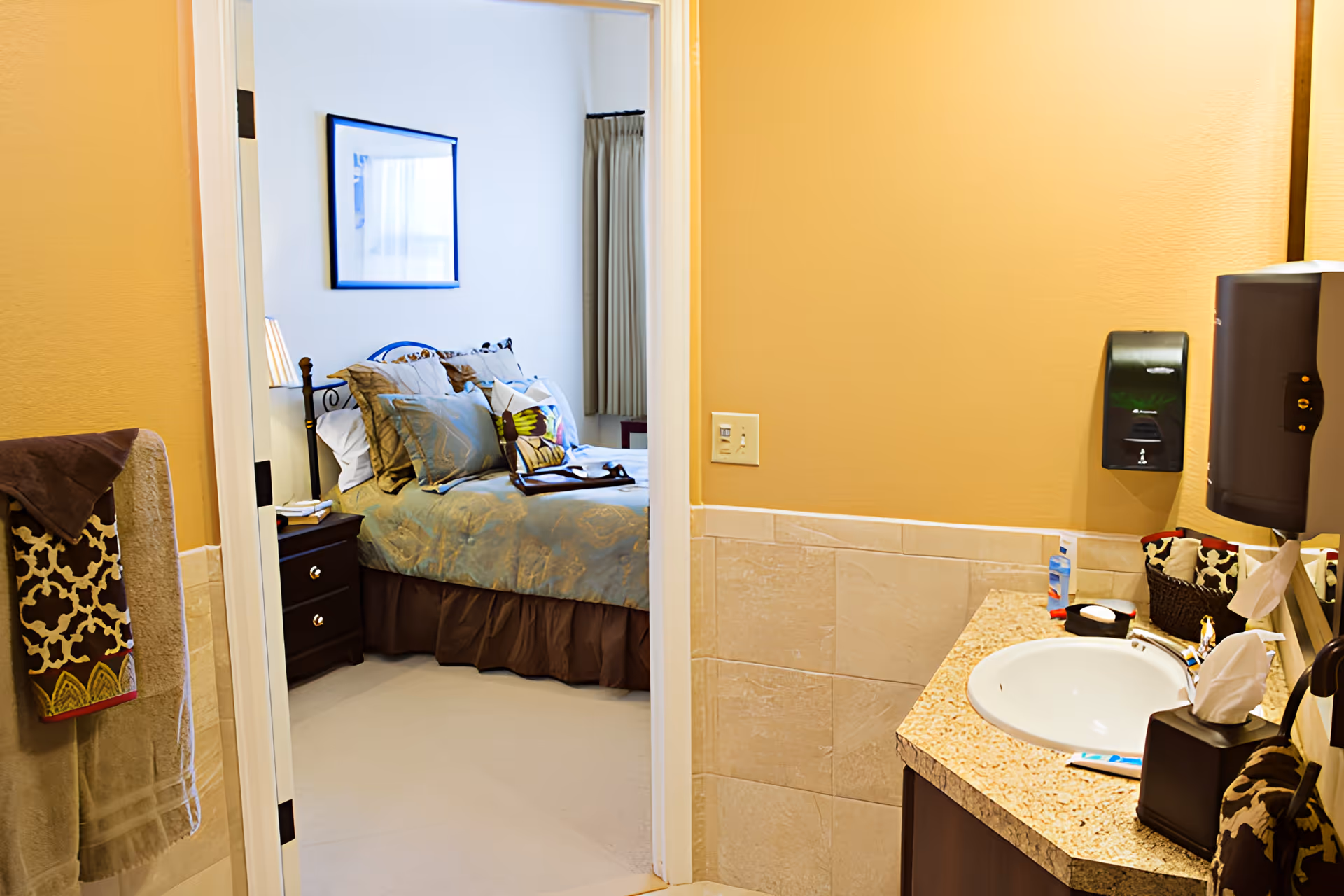 View from a bathroom into a bedroom in a senior living facility. The bathroom has a granite countertop with a white sink, a soap dispenser, tissue box, and towels hanging on the wall. The bedroom features a neatly made bed with multiple pillows, a nightstand with a lamp, and a framed picture on the wall.