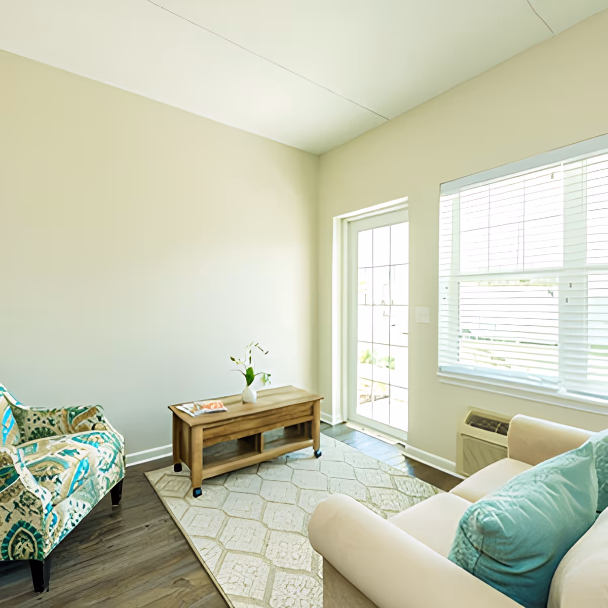 Bright small living room with a sofa and patterned armchair around a wooden coffee table near a windowed door.