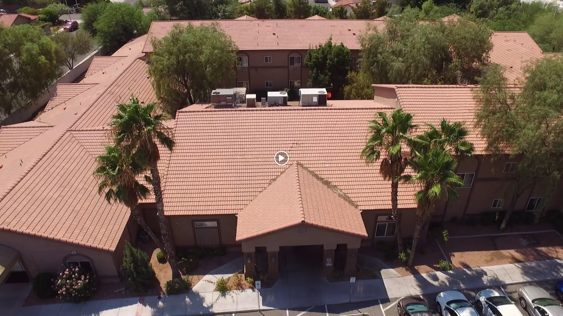 Aerial view of Sana Assisted Living facility showing a large building with a red-tiled roof, surrounded by trees and palm trees. There is a covered entrance at the front with a driveway and several parked cars visible. The building is set in a residential area with greenery around.