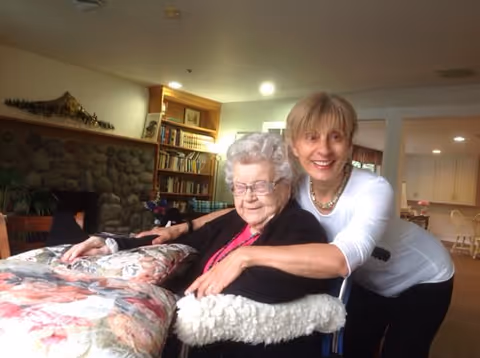 An elderly woman sitting in a wheelchair covered with a floral blanket, smiling, with a younger woman standing behind her and hugging her. They are in a cozy room with a stone fireplace, bookshelves, and warm lighting.