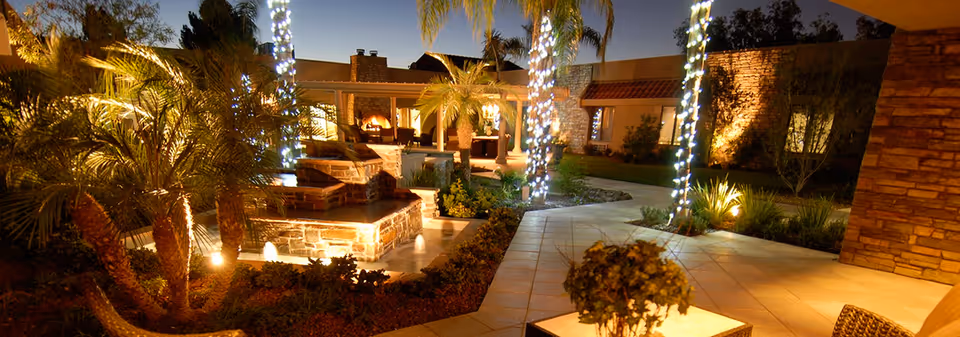 A nighttime outdoor courtyard with palm trees wrapped in string lights, a stone fountain, tiled walkways, and a covered seating area.