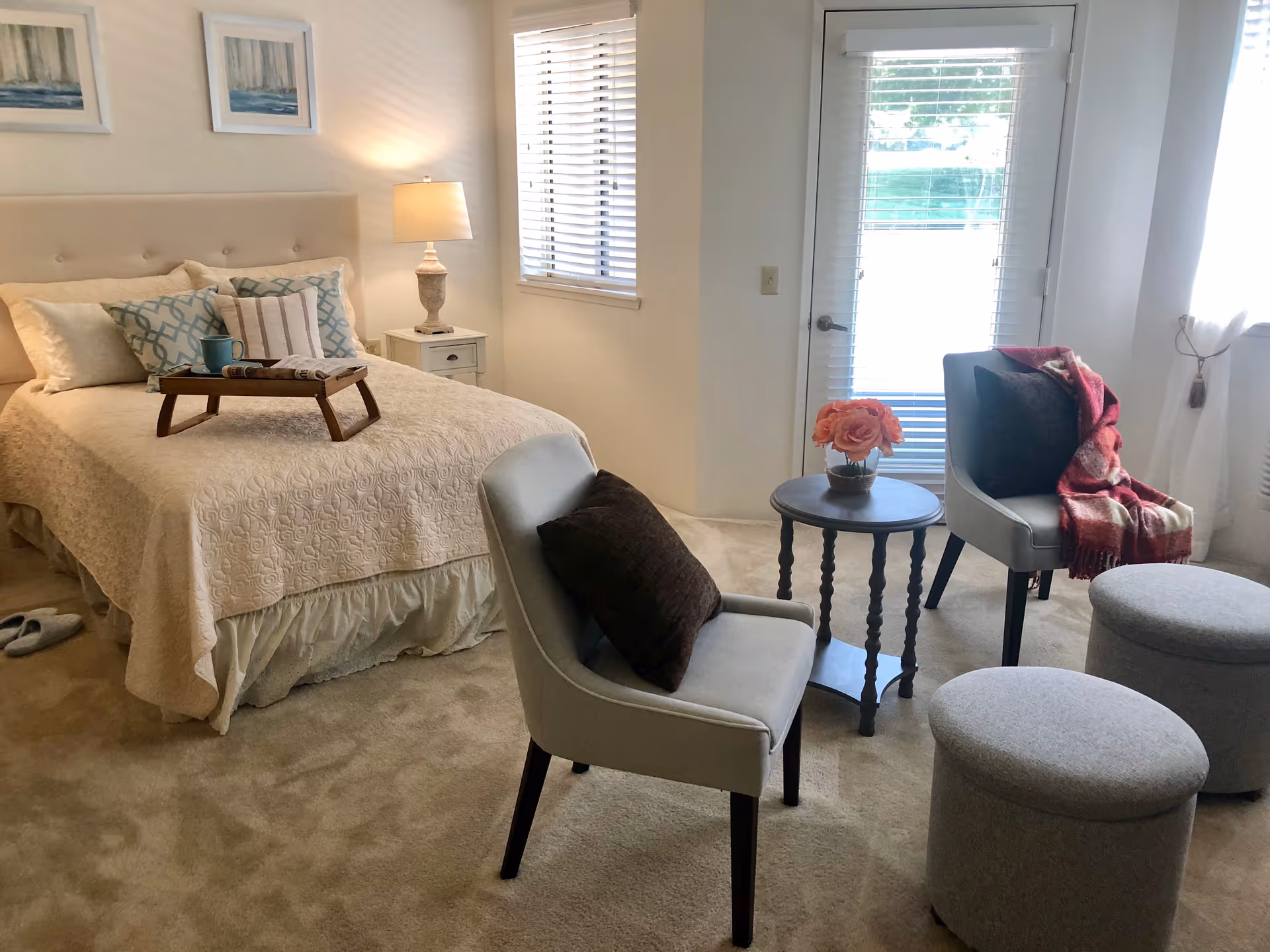 A cozy bedroom with a neatly made bed featuring a beige quilt and multiple pillows. A wooden tray with a cup and a book rests on the bed. Next to the bed is a nightstand with a lamp. Two upholstered chairs with dark cushions and two round ottomans surround a small round table with a vase of pink flowers. The room has carpeted flooring, a window with blinds, and a glass door leading outside.