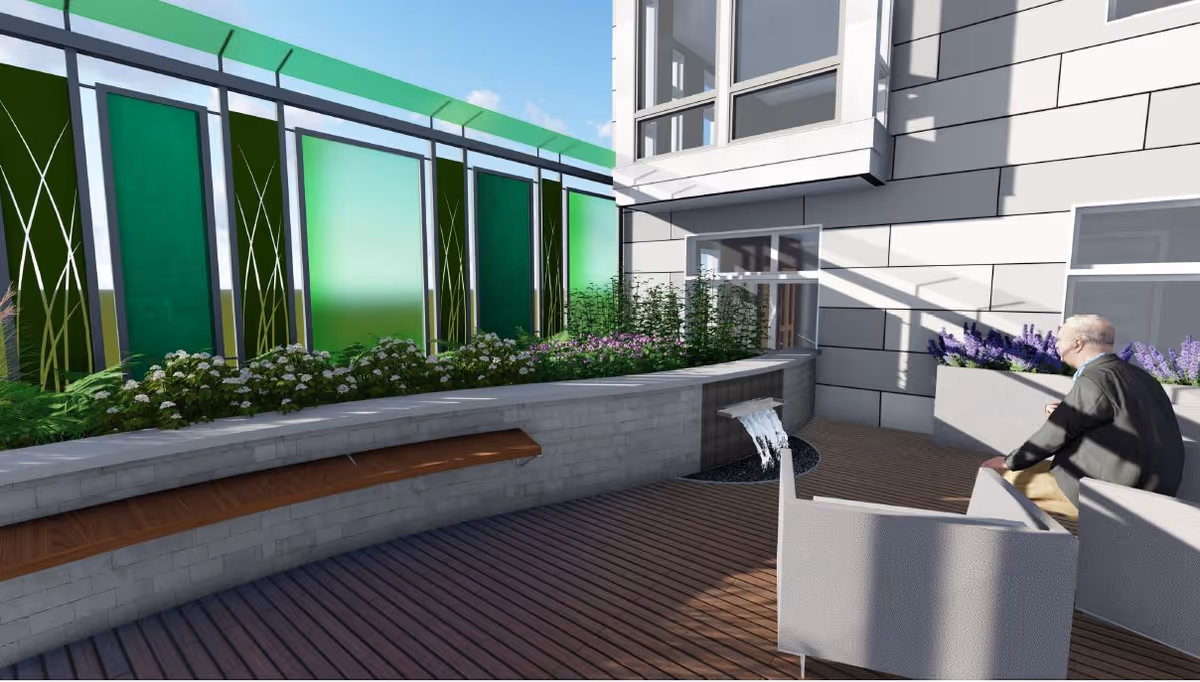A sunny outdoor courtyard with planter beds, green decorative panels, a small water feature, benches and chairs, and an elderly man seated near the building.