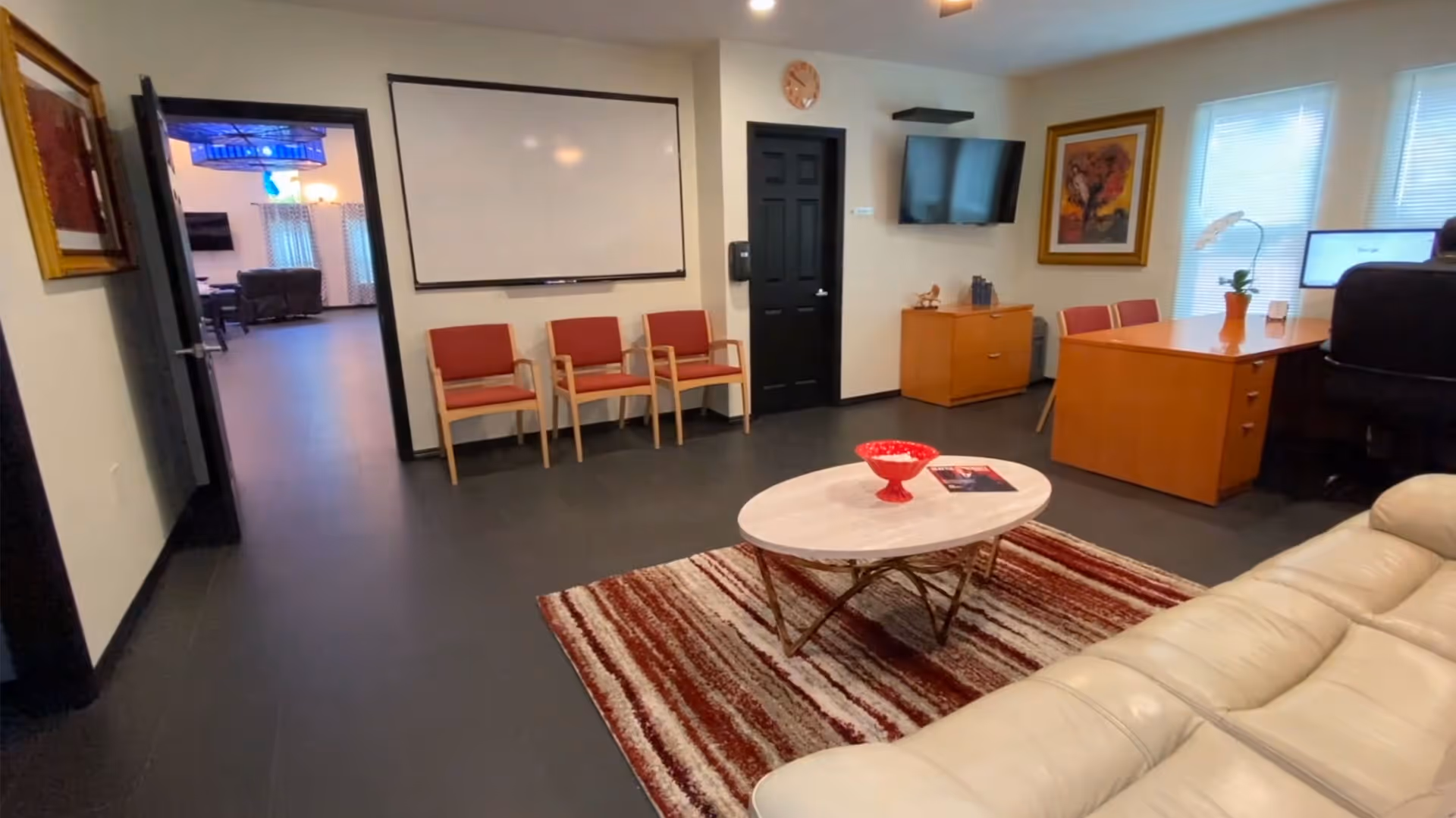 Small reception/lounge interior with a sofa and coffee table on a rug, red chairs along the wall and a desk with a mounted TV.