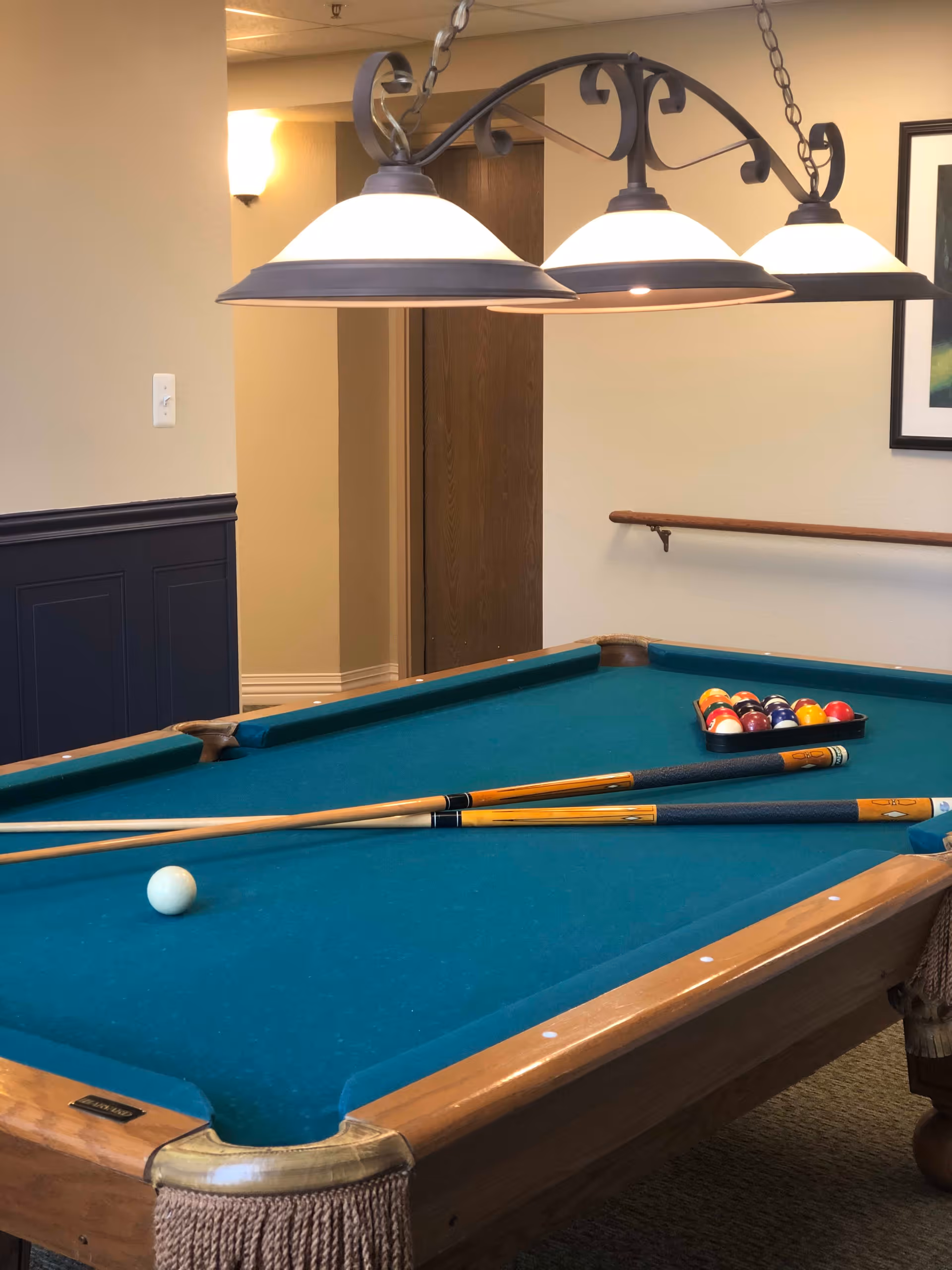 A pool table with a green felt surface, two pool cues resting on it, and a rack of billiard balls arranged at one end. Above the table, three hanging lamps provide lighting. The room has beige walls, a wooden door, a handrail on the wall, and a framed picture partially visible on the right.