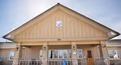 Exterior front view of a single-story building with a peaked roof and beige siding. The building has a covered entrance with railings and two wall-mounted lantern-style lights. The number 209 is displayed above the entrance, and there is a small sign with a logo near the roof peak. The sky is clear and blue.