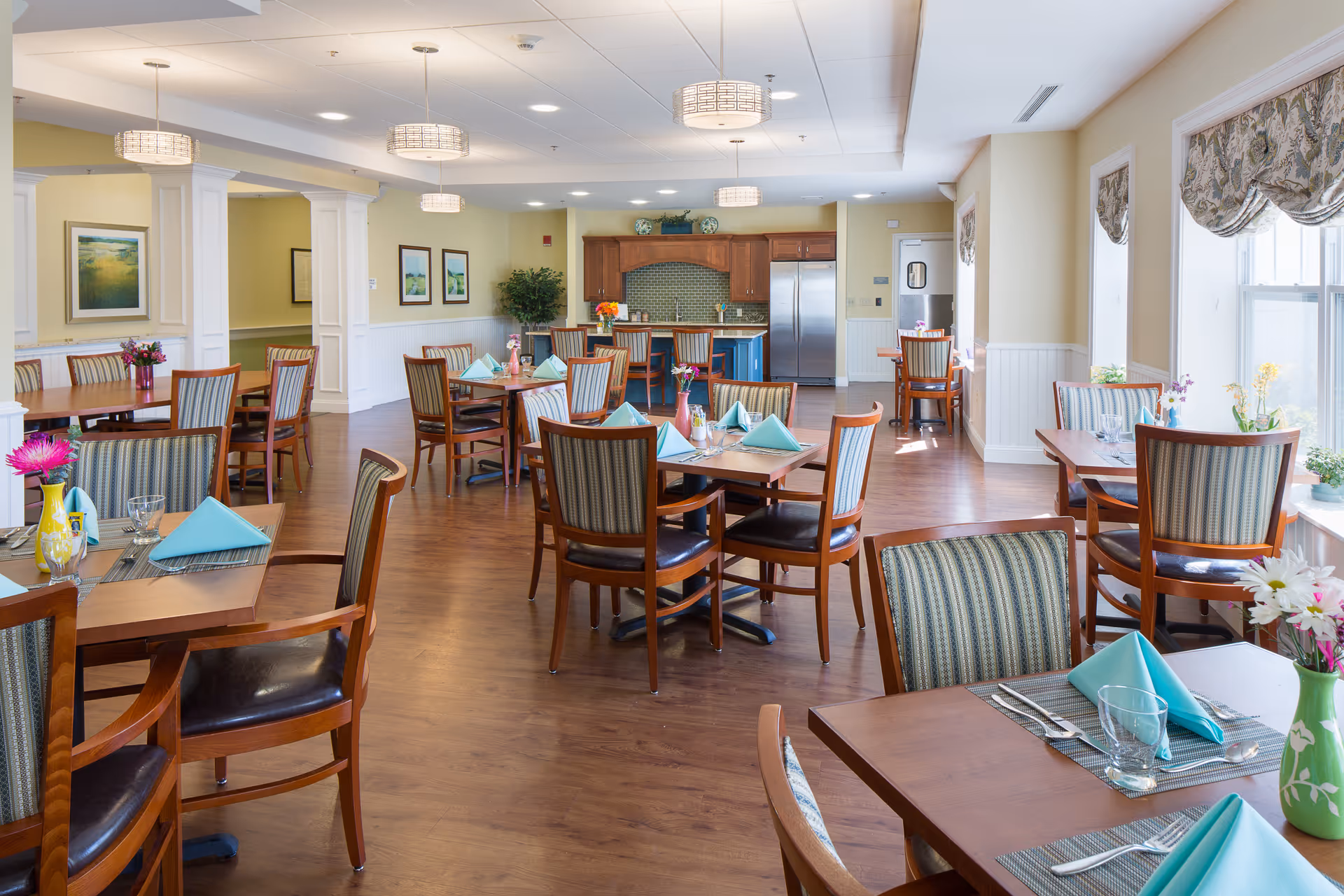 A bright and spacious dining room with wooden tables and chairs arranged neatly. Each table is set with turquoise napkins, glassware, and cutlery. The room features large windows with patterned valances allowing natural light to fill the space. There are pendant lights hanging from the ceiling and a kitchen area with stainless steel appliances and wooden cabinetry in the background.