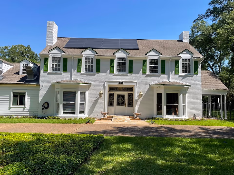 Two-story white brick house with green shutters, solar panels on the roof and a central entrance set in a landscaped lawn.