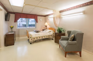 A well-lit patient room in a healthcare facility featuring a single hospital bed with a patterned quilt, a bedside table with a lamp and a plant, a comfortable armchair with a pillow, a small dresser with a vase of flowers, a wall-mounted TV, and a window with a red valance showing an outdoor view.