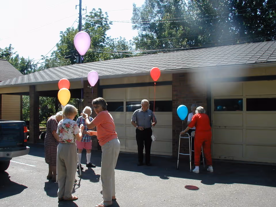 A group of elderly people standing outside near a garage, each holding colorful balloons. Some are using walkers for support. The scene is sunny with trees in the background.