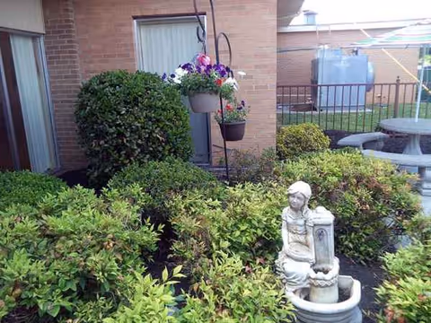 Outdoor courtyard with bushes, hanging planters, and a statue fountain beside a brick building.
