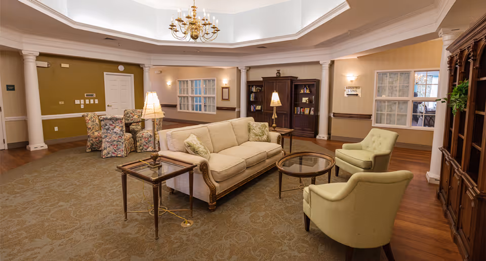 A spacious living room area with a beige sofa, two light green armchairs, a glass coffee table, and side tables with lamps. There are bookshelves against the walls, floral-patterned chairs around a small table, and a chandelier hanging from the ceiling. The room has warm lighting, carpeted floors, and large windows.