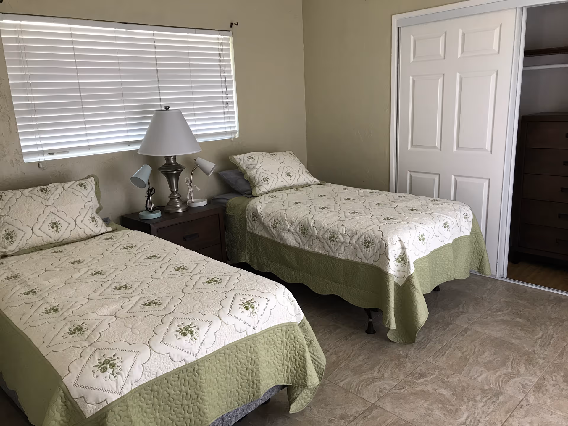 A bedroom with two twin beds covered in matching green and white floral quilts. Between the beds is a dark wooden nightstand with a large lamp and two smaller lamps. A window with closed white blinds is above the nightstand. There is a closet with sliding white doors and a dresser inside the closet.