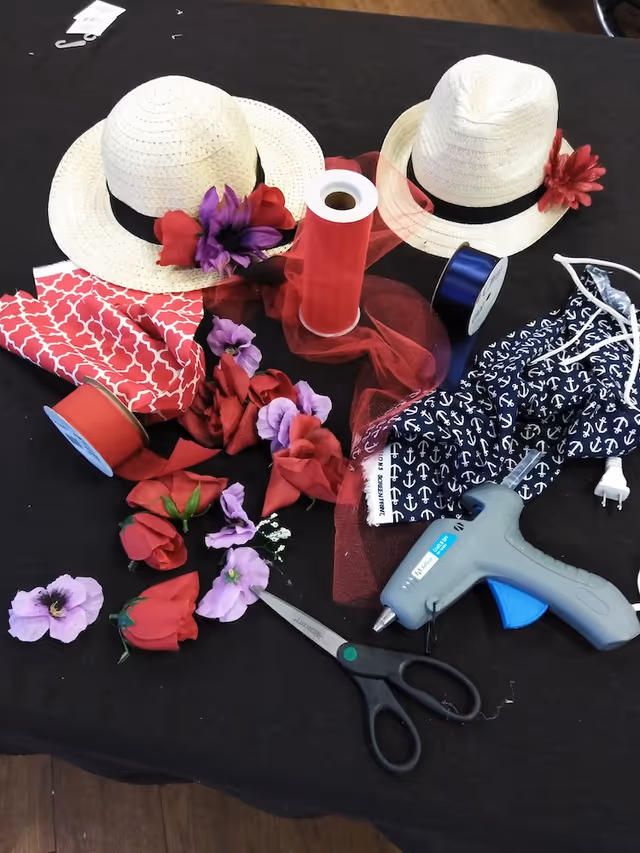 A crafting setup on a black surface featuring two straw hats decorated with artificial red and purple flowers, various red and purple artificial flowers scattered around, rolls of red and blue ribbon, a pair of scissors, a hot glue gun, and a piece of navy blue fabric with white anchor patterns.