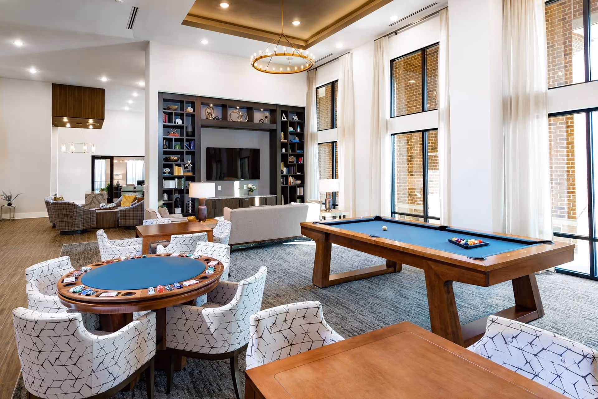 Spacious bright communal lounge featuring a poker table, pool table, seating area, bookshelves, and tall windows with sheer curtains.
