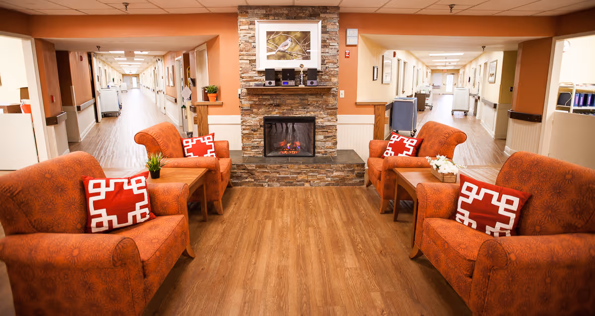 A cozy seating area in a senior living facility with four orange upholstered armchairs arranged around two wooden side tables. Each chair has a red and white decorative pillow. In the center background, there is a stone fireplace with a framed picture of a bird above it. The room has wood flooring and two long hallways extending on either side of the fireplace.