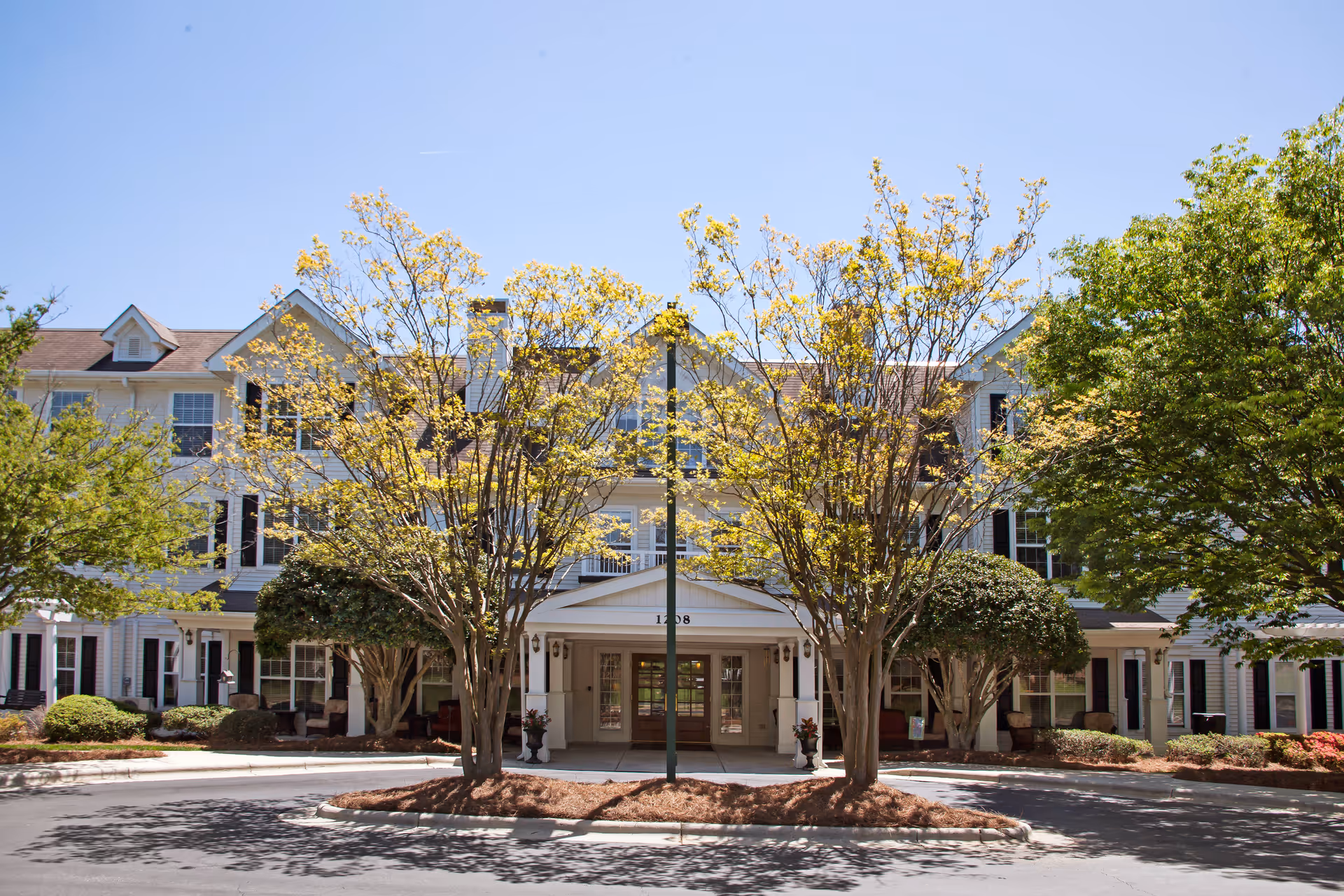 Front façade of a three-story senior living building with trees framing the covered entrance and a circular driveway.