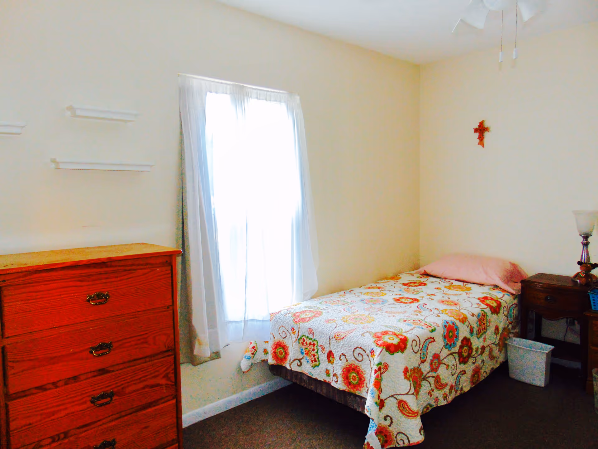 Small bedroom with a single bed covered in a floral quilt, a wooden dresser, nightstand and a window with sheer curtains.