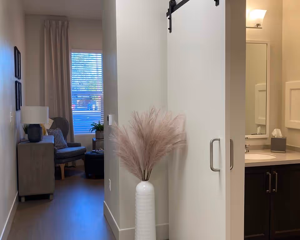 View down a hallway in an assisted living facility showing a white sliding barn door partially open to reveal a bathroom sink and mirror. A white vase with tall, fluffy pampas grass is placed against the wall near the door. In the background, there is a sitting area with a gray armchair, a small table with a lamp, and a window with blinds and curtains.