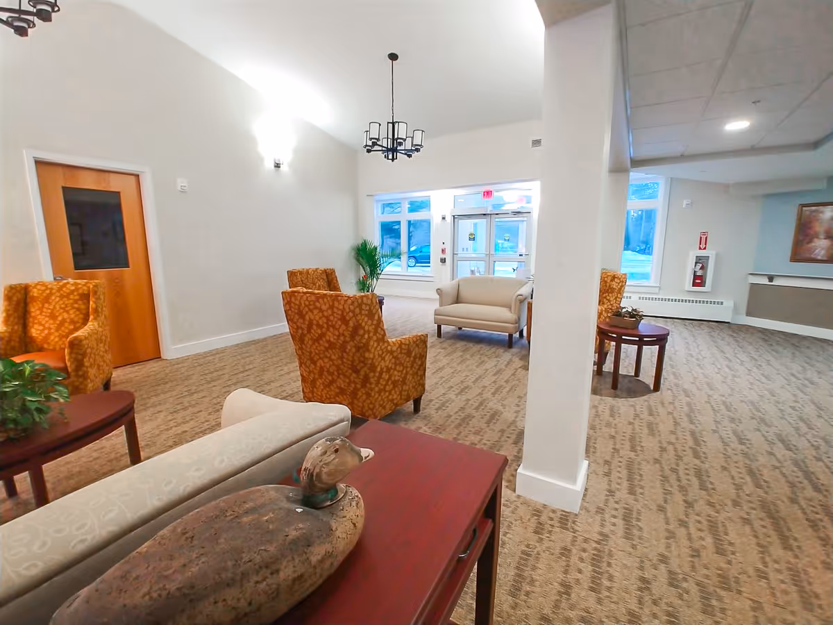 A spacious and well-lit common area in a senior living facility featuring patterned carpet, several upholstered chairs in orange and beige tones, a small beige loveseat, a wooden table with a decorative duck sculpture, potted plants, and large windows near the entrance door allowing natural light to enter.