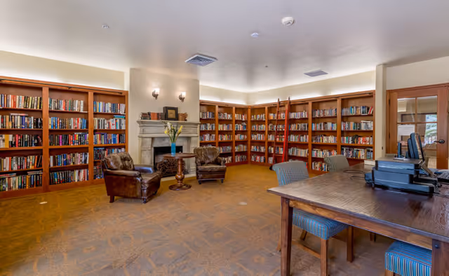 Spacious senior living library with built-in bookshelves, leather armchairs by a fireplace, and a wooden table with chairs and computers.