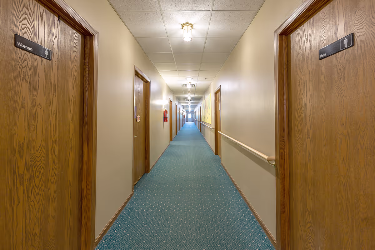 Long hallway in a senior living facility with beige walls, green carpet, and wooden doors on both sides. The doors on the left and right closest to the camera are labeled Women and Men, indicating restrooms. Ceiling lights illuminate the corridor, and handrails run along the walls.