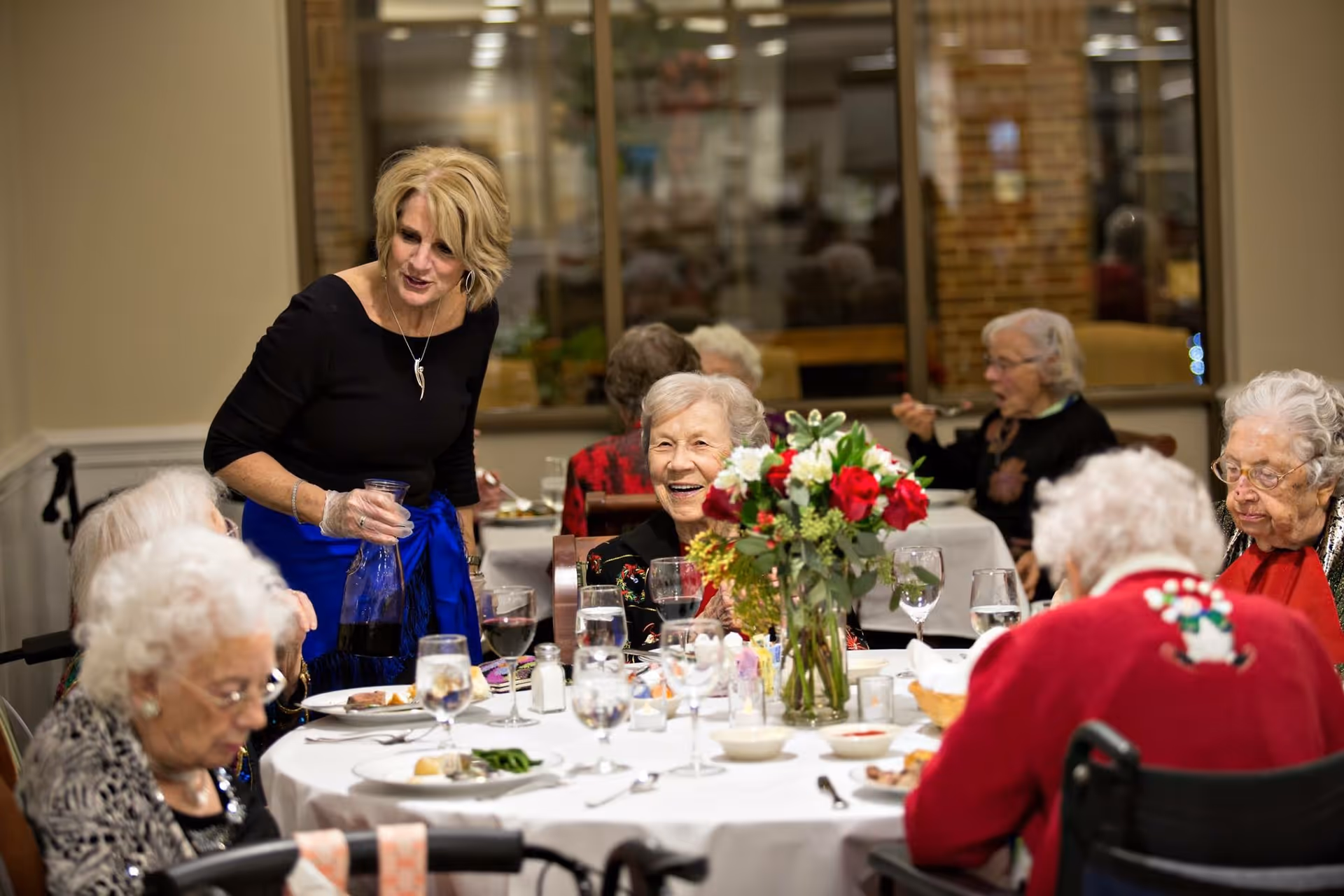 A group of elderly women seated around a dining table with white tablecloth, enjoying a meal together. A woman standing is pouring a drink for one of the seated women. The table is set with plates, glasses, and a floral centerpiece with red and white flowers. The setting appears to be a communal dining room with large windows in the background.