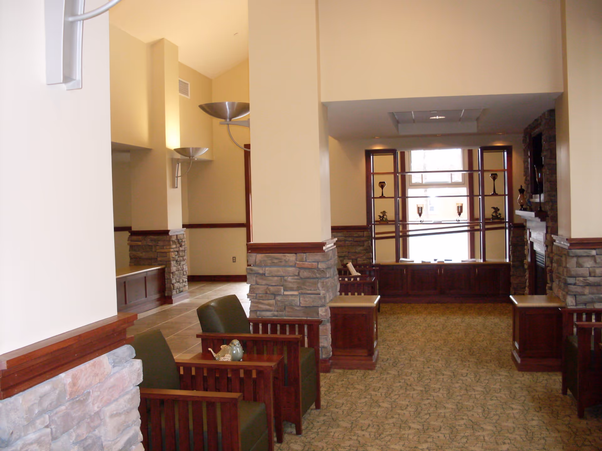 Interior view of a senior living facility lounge area with stone pillars, wooden furniture including green cushioned chairs, a carpeted floor, and a large window with decorative shelves. The walls are painted beige with wooden trim and there are modern wall-mounted light fixtures.