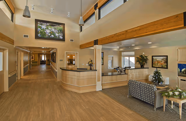 Interior view of Columbine Commons Assisted Living facility showing a spacious reception area with a curved wooden front desk, a seating area with a patterned sofa and a small decorated Christmas tree, wooden beams on the ceiling, pendant lights, and a hallway leading to other rooms. The walls are adorned with framed artwork and there are large windows allowing natural light.