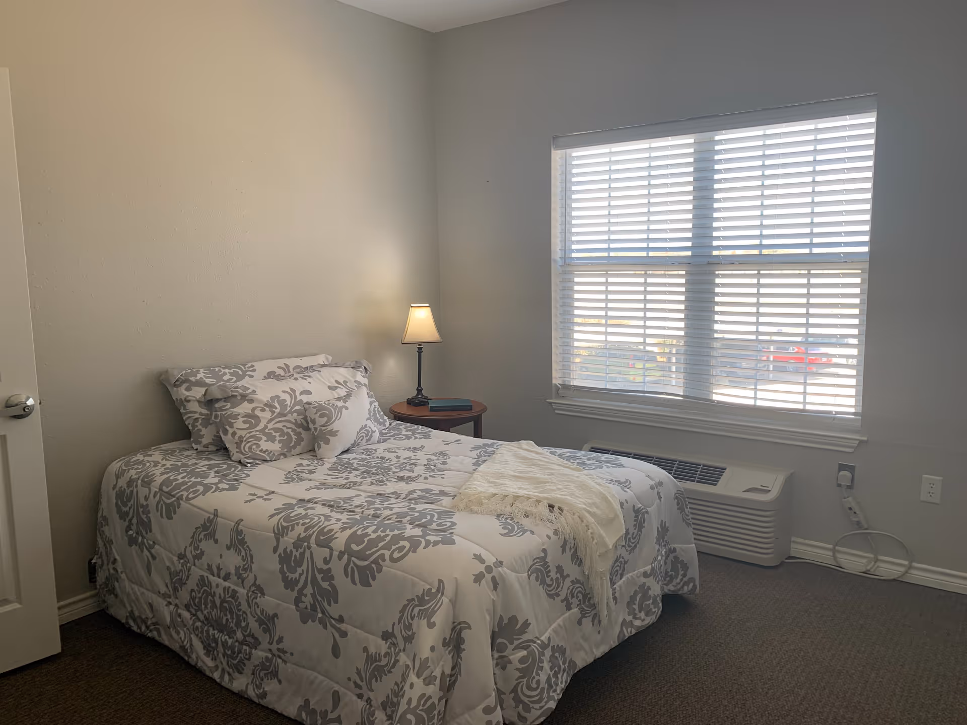 Small bedroom with a single bed dressed in patterned bedding, a bedside table with a lamp, and a window with blinds.