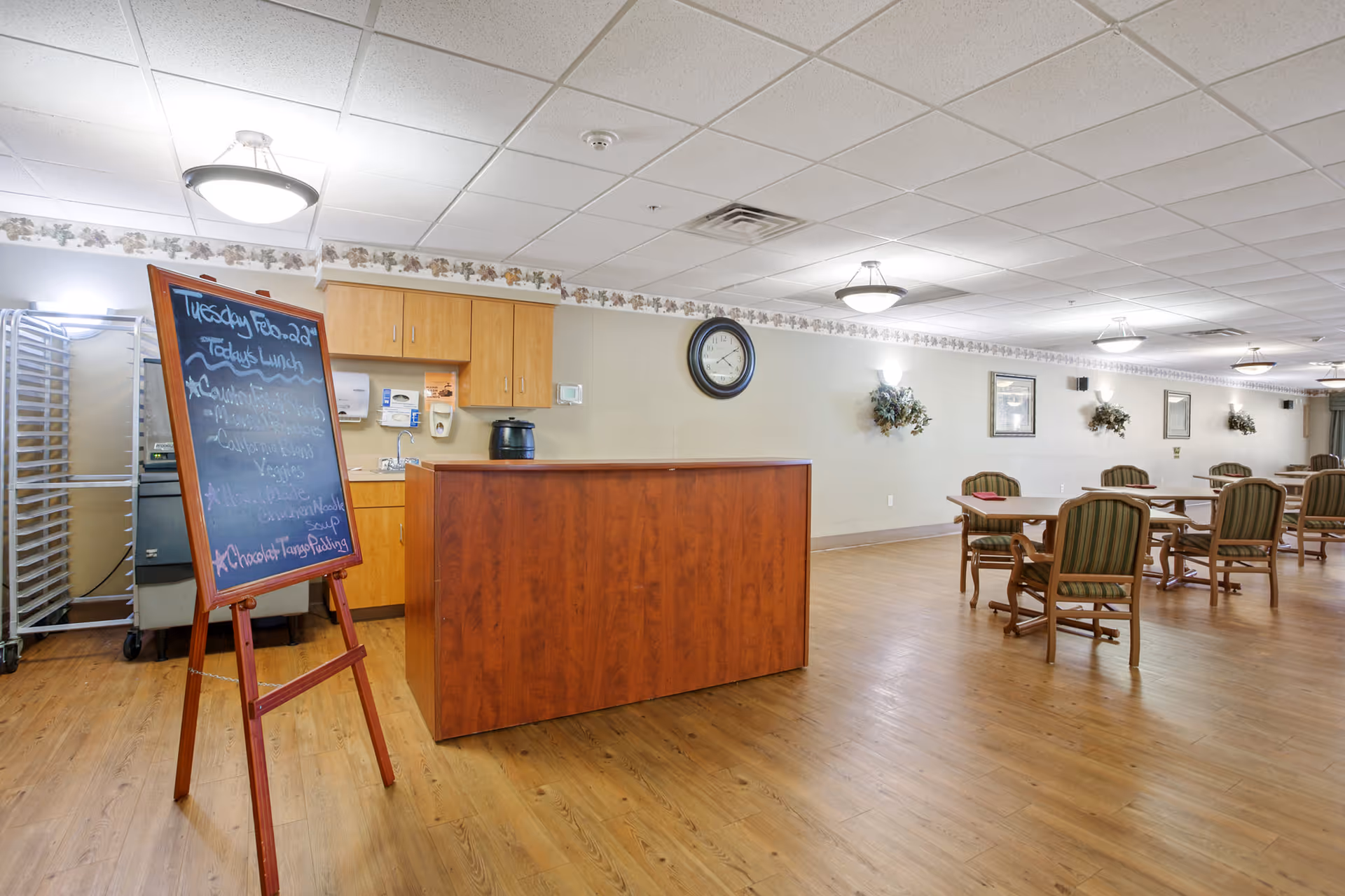 Interior view of a dining area in a senior living facility with wooden floors, several tables and chairs, a wooden counter, and a chalkboard displaying the lunch menu. The room is well-lit with ceiling lights and decorated with wall-mounted plants and framed pictures.