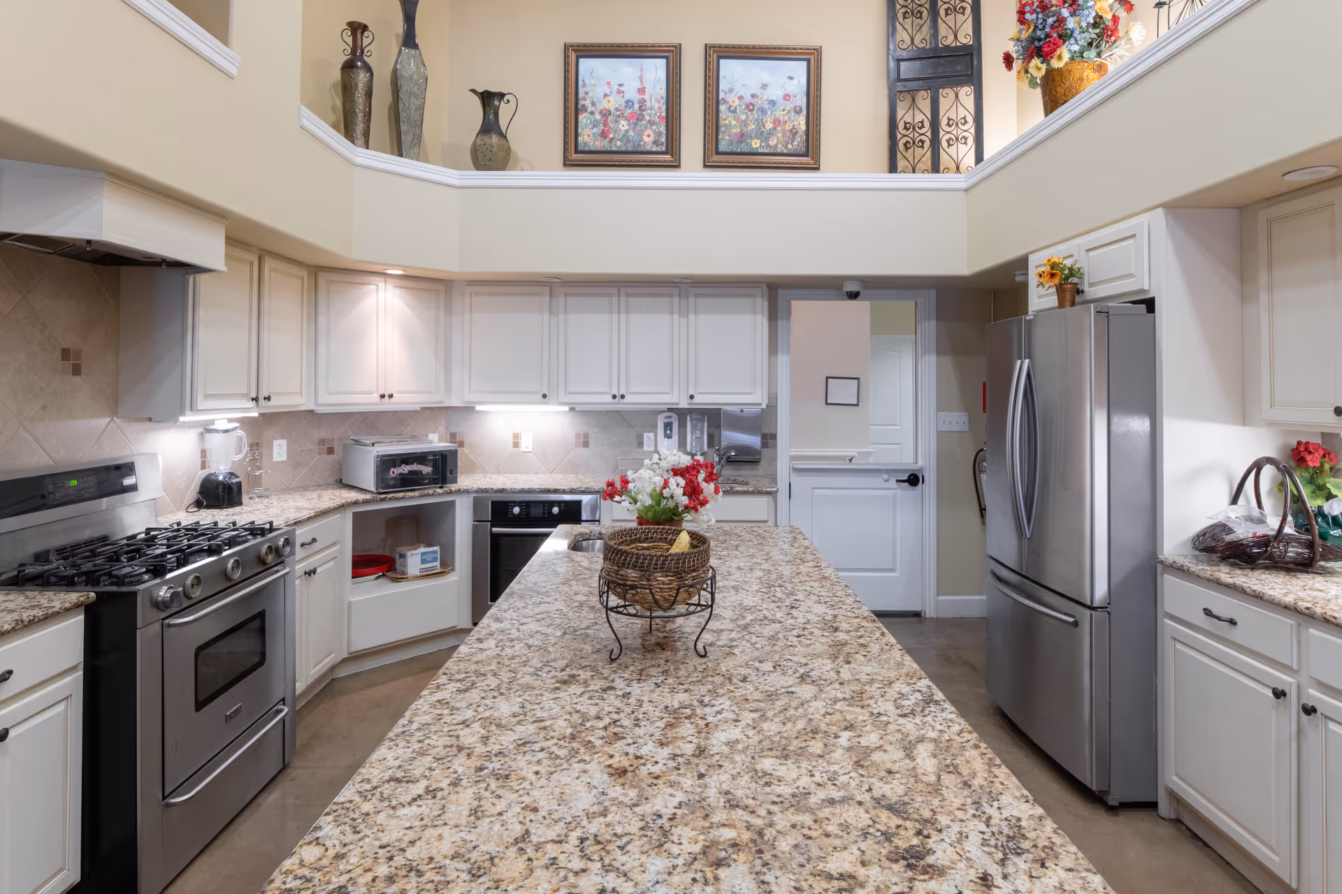 A spacious kitchen with granite countertops and white cabinetry. The kitchen features a large central island with a decorative basket and flowers, stainless steel appliances including a refrigerator, oven, and gas stove. The walls are painted beige, and there are decorative vases and framed floral artwork on a ledge above the cabinets. The kitchen is well-lit with under-cabinet lighting and recessed ceiling lights.