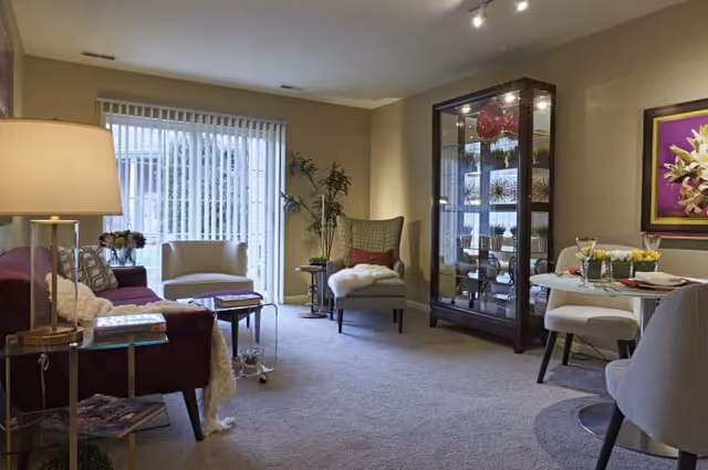 A cozy living room area with a purple sofa, a beige armchair, a glass display cabinet filled with decorative items, and a small dining table set for two. The room has beige walls, carpeted floor, a large window with vertical blinds, and soft lighting from a table lamp and ceiling lights.