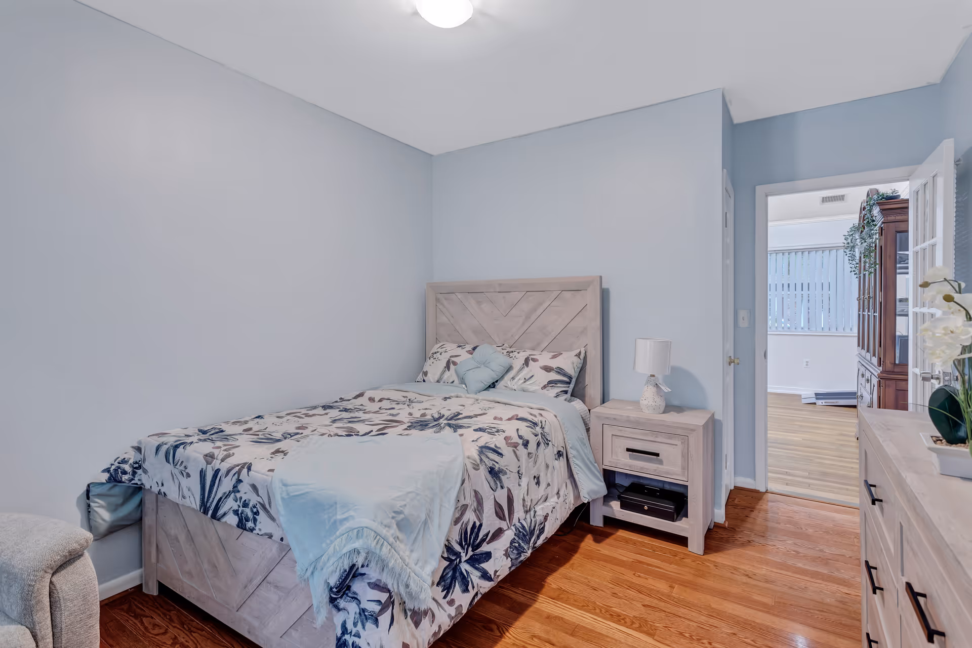 A bedroom with light blue walls and wooden floor. The room features a bed with a floral patterned comforter and light blue pillows, a matching wooden nightstand with a white lamp, and a dresser with decorative items. An open door leads to another room with wooden flooring and a large window with vertical blinds.