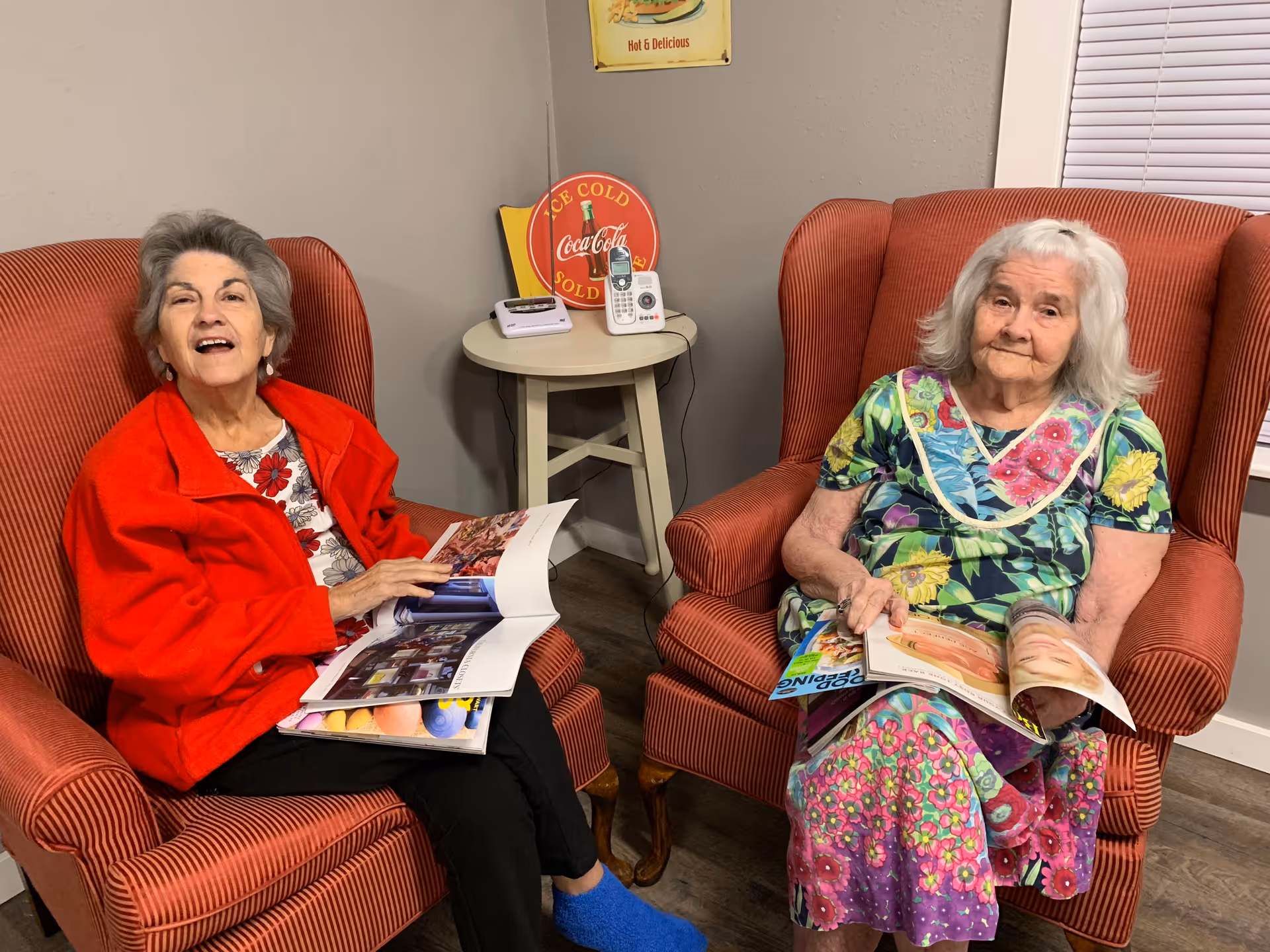 Two elderly women sitting in red armchairs in a cozy room. One woman is wearing a red jacket and black pants, smiling while holding a magazine. The other woman is wearing a colorful floral dress, looking at the camera while also holding a magazine. A small round table with a cordless phone and a Coca-Cola sign is between them. The room has gray walls and a window with blinds.